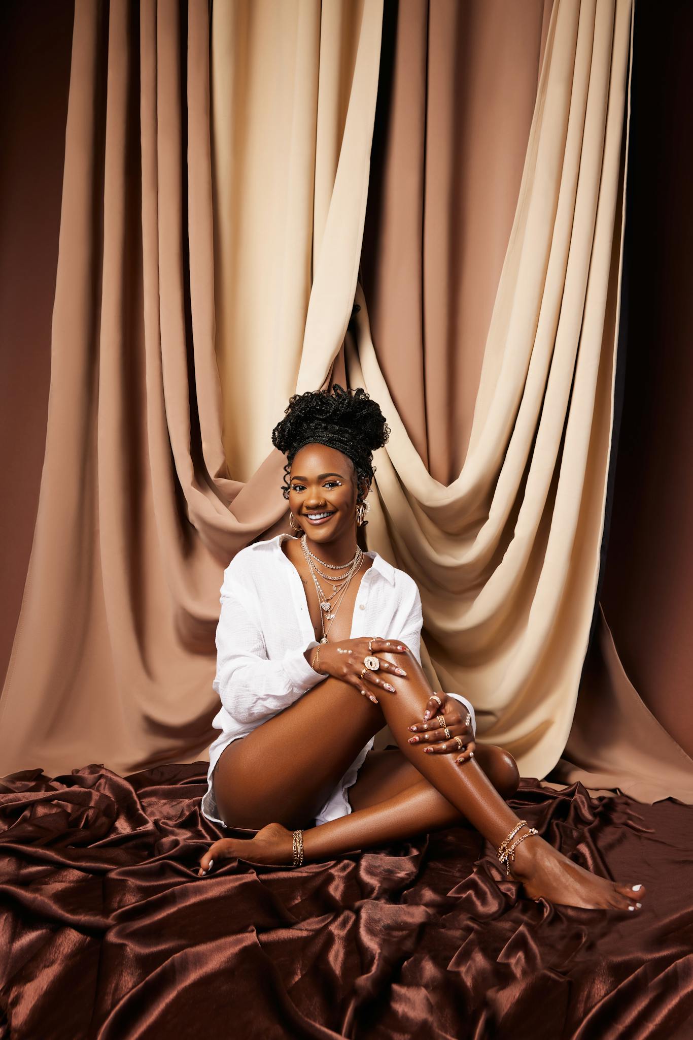 Portrait of a smiling woman seated amidst luxurious brown fabrics in a studio setting.