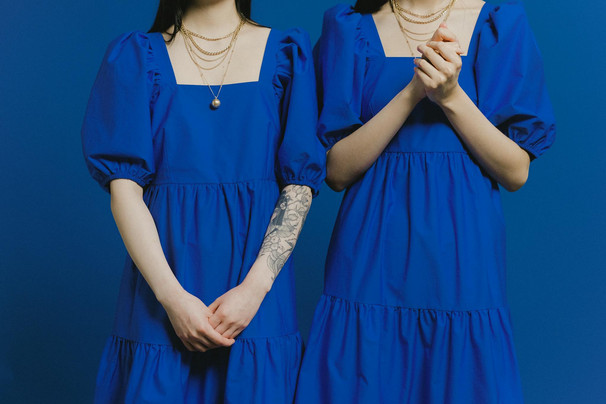 Two women in vibrant blue dresses, adorned with gold necklaces, against a blue background.